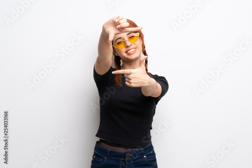 Young redhead woman over white wall focusing face. Framing symbol