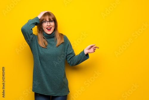 Woman with glasses over yellow wall pointing finger to the side and presenting a product