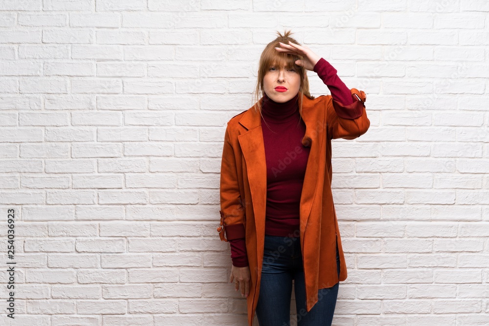 Woman with coat over white brick wall looking far away with hand to look something Woman with coat over white brick wall looking far away with hand to look something