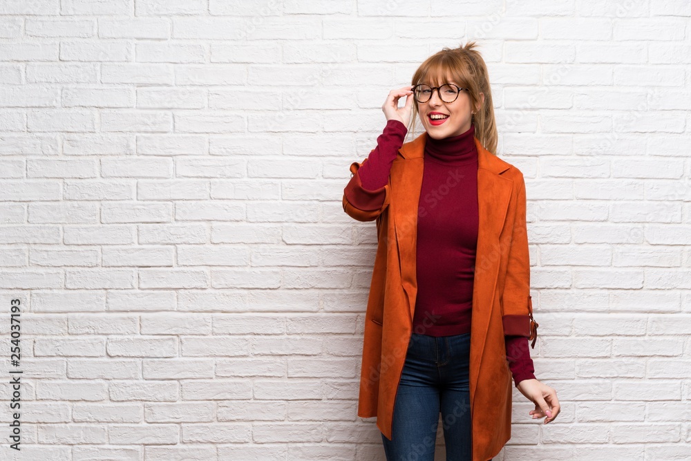 Woman with coat over white brick wall with glasses and surprised Woman with coat over white brick wall with glasses and surprised