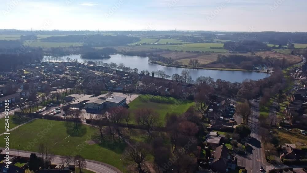 Ascending aerial view across rural British countryside town overlooking lake along green country farming pastures & hazy meadows in bright sunshine. Ascending aerial view across rural British countryside town overlooking lake along green country farming pastures & hazy meadows in bright sunshine.
