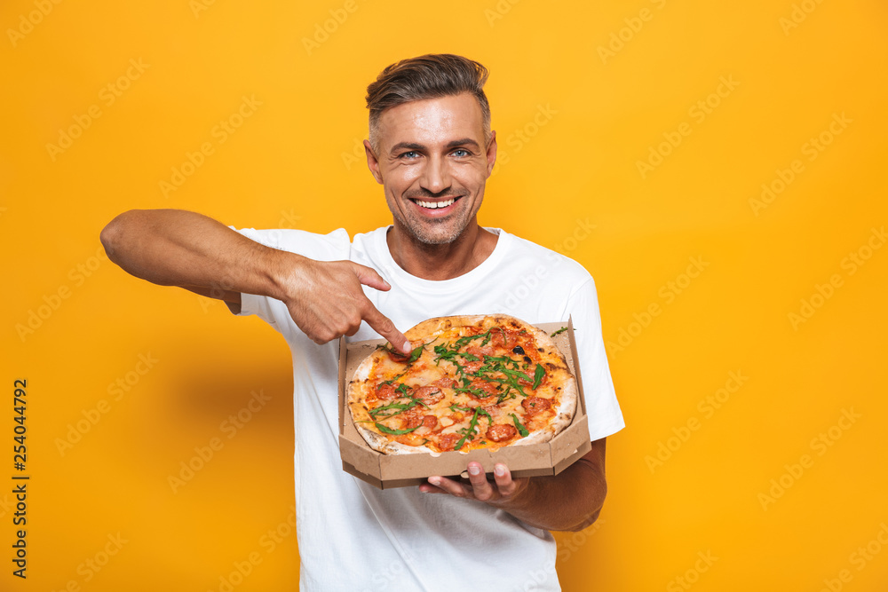 Image of optimistic man 30s in white t-shirt holding and eating pizza while standing isolated