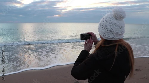 Adult woman taking photos of the seascape using a mobile phone on a sandy beach. Autumn mood.