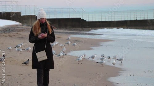 Adult woman walks along a sandy beach with a mobile phone among a flock of gulls. Autumn mood.
