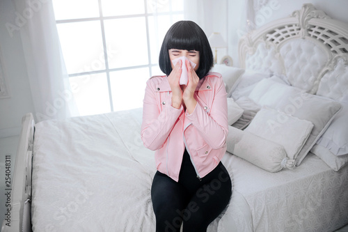 Fashionable freak. Glamour woman with short black hair is blowing nose and suffering from ill in the white bedroom. Stylish girl in pink jacket. Fashion and health care concept
