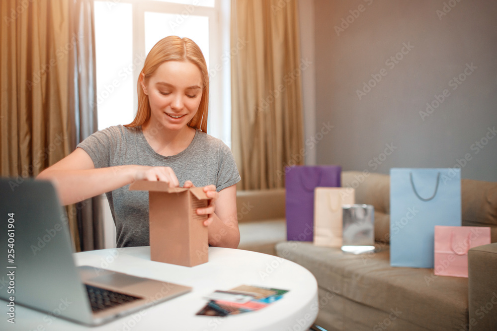Online shopping at home. Young smiling shopper is unboxing her parcel, ordered by internet, while sitting at the table Online shopping at home. Young smiling shopper is unboxing her parcel, ordered by internet, while sitting at the table