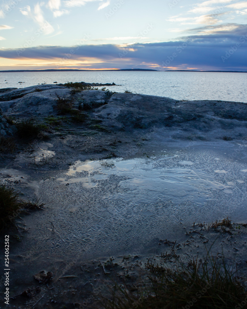 Obraz premium Ice in an interesting pattern on rocks overlooking the ocean, with an island in the distance and colorful sky during a sunset. Intentionally dark to create a moody photograph.