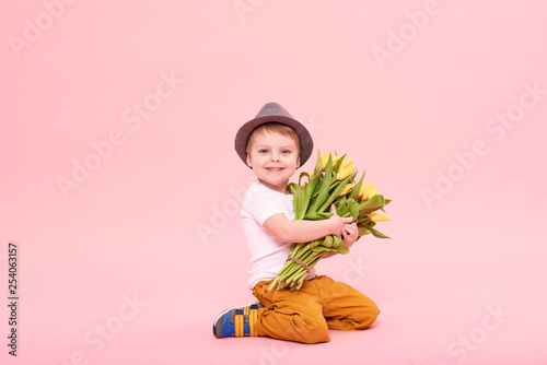 Adorable smiling child with spring flower bouquet looking at camera isolated on pink. Little toddler boy in hat holding yellow tulips as gift for mom. Copy space for text  #254063157
