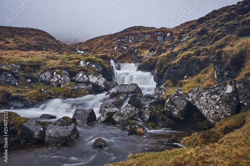 Wild stream or creek in moorland or heath in the highland of Faroe island Vagar with waterfall cascades and stones with moss and lichen on them. Picture taken in cloudy, misty, foggy and rainy day.