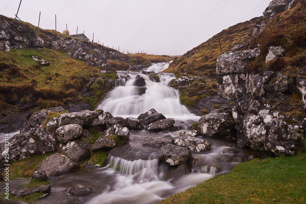 Wild creek or stream with waterfalls and water cascade between fences of sheep pasture or grazing land during the cold, foggy, místy and rainy day in spring on Faroe islands in north atlantic ocean. Wild creek or stream with waterfalls and water cascade between fences of sheep pasture or grazing land during the cold, foggy, místy and rainy day in spring on Faroe islands in north atlantic ocean.