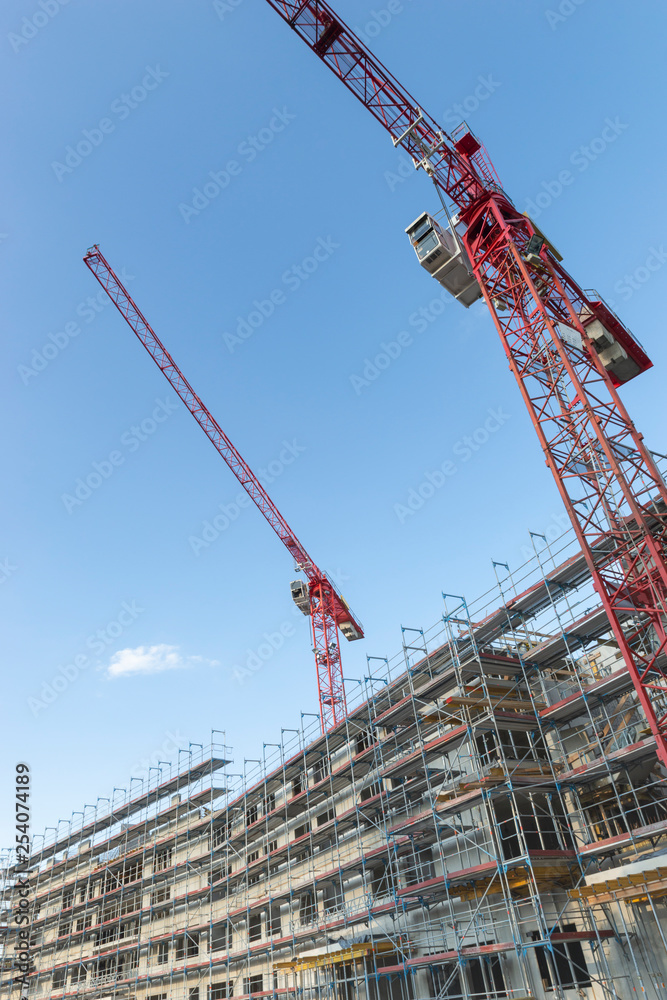 In blauen Himmel aufragende Baukräne, die um einen eingerüsteten Rohbau stehen. In blauen Himmel aufragende Baukräne, die um einen eingerüsteten Rohbau stehen.