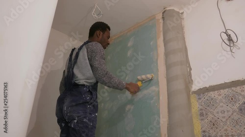 black guy works on a construction site, paints the walls with glue using a rolle