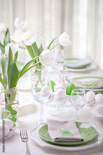 Decor and table setting of the Easter table with white tulips and dishes of green and white color. Easter decor in the form of Easter bunnies green color with white polka dots.