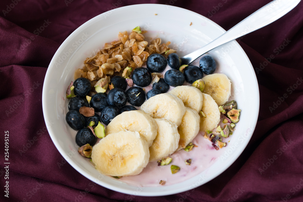 Bowl of fresh blueberries, banana and muesli yogurt on red color table clothes
