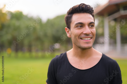 Face of happy young handsome Hispanic man thinking at the park