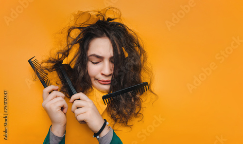 One comb in the hair and two in the hands do not help to cope with the problem.A funny teenage caucasian girl with problematic dry curly hair expresses displeasure on her face.Orange paper background.