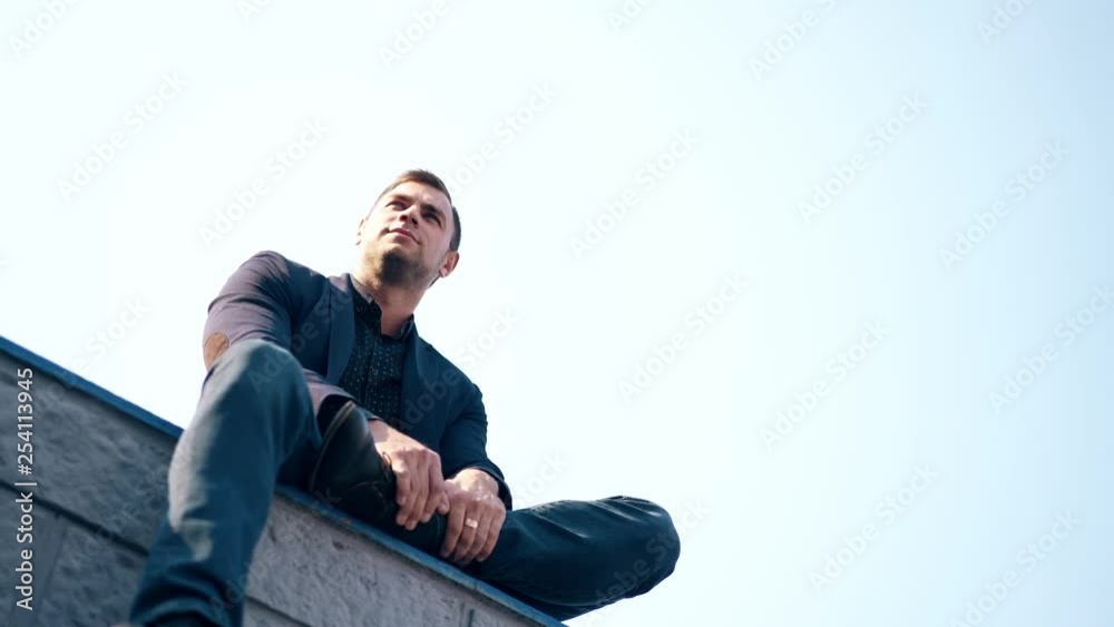 low angle shot guy in suit sits on parapet and fixes black hair by hand with golden ring under white summer sky low angle shot guy in suit sits on parapet and fixes black hair by hand with golden ring under white summer sky