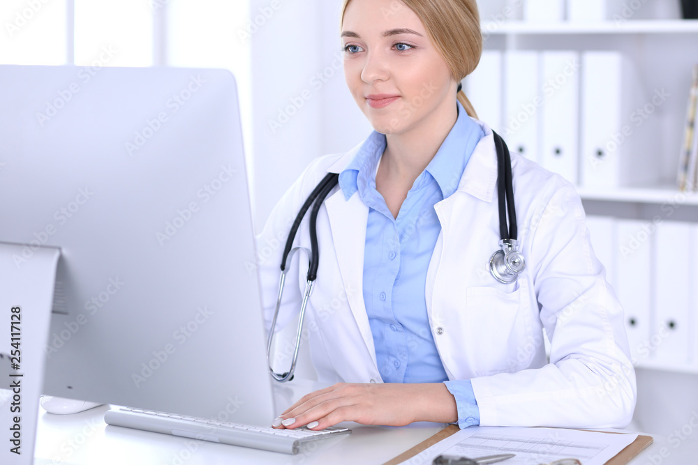 Young woman doctor at work in hospital looking at desktop pc monitor. Physician controls medication history records and exam results. Medicine and healthcare concept
