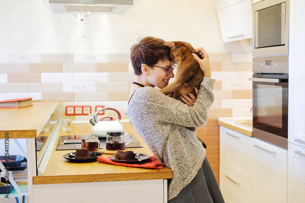 Smiling woman playing with a ginger cat, hugging and fooling around in the kitchen with him. The mood of love and laughter. Smiling woman playing with a ginger cat, hugging and fooling around in the kitchen with him. The mood of love and laughter.
