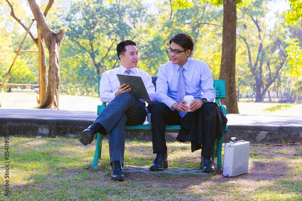 Fototapeta premium Businessman, They are sitting on bench in park. He is play notebook and search internet. Another one holding paper and talking about business in relax time. Photo concept business and relax time.