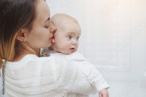 Young happy loving mother plays kisses her baby daughter in her arms against the window. Maternal care. Childcare. Close-up.