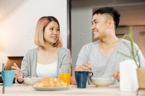happy asian couple in pajamas sitting at table in kitchen at home in morning and having cereal breakfast together.