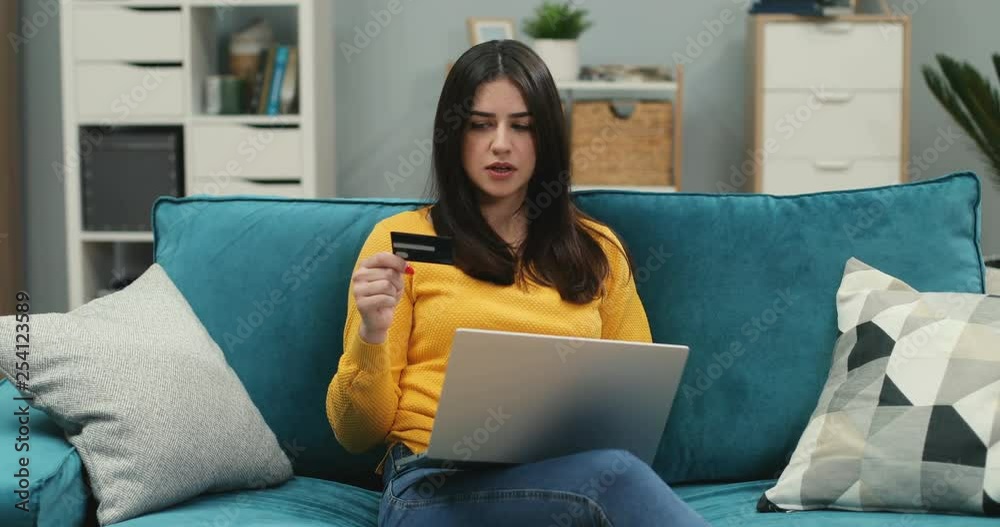Young Caucasian woman sitting on the sofa in the living room and shopping online with a credit card on the laptop computer, then being very cheerful from the successful bought. Young Caucasian woman sitting on the sofa in the living room and shopping online with a credit card on the laptop computer, then being very cheerful from the successful bought.