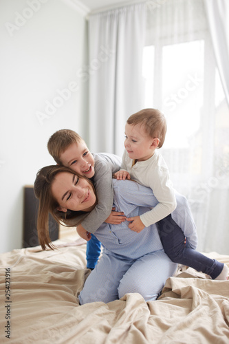 Beautiful young mother and her two little sons dressed in pajamas have fun on the bed in the light cozy bedroom