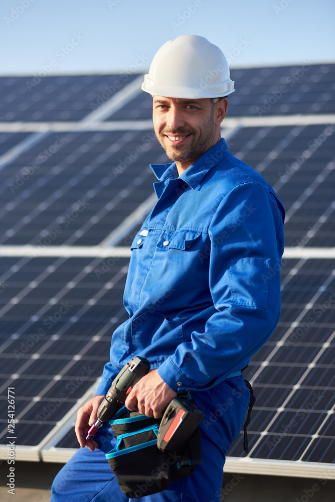 Portrait of male worker in blue suit and protective helmet installing solar photovoltaic panel system. Electrician smiling to the camera and standing on roof of house. Alternative energy concept. Portrait of male worker in blue suit and protective helmet installing solar photovoltaic panel system. Electrician smiling to the camera and standing on roof of house. Alternative energy concept.