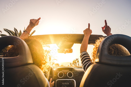 Couple of woman friends traveling and driving having a lot of fun dancing in the car with opened roof and summer vacation sunset ocean in front - concept of friendship together and nice lifestyle