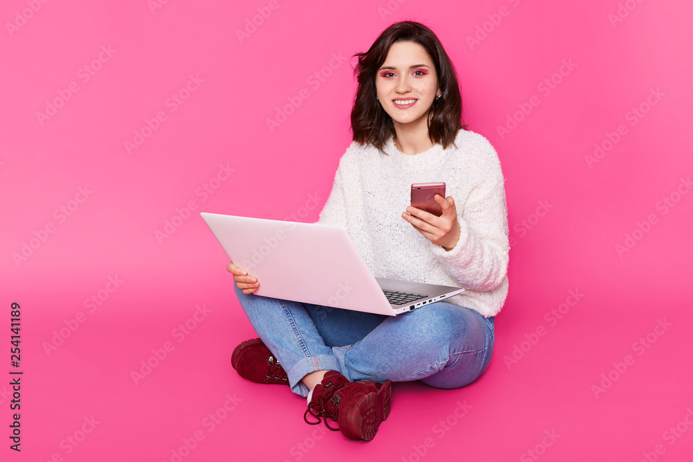 Portrait of smiling young woman wears white sweater, fashion jeans and claret shoes. Charming girl sits with crossed legs, holds laptop computer and modern smathphone isolated over pink background. Portrait of smiling young woman wears white sweater, fashion jeans and claret shoes. Charming girl sits with crossed legs, holds laptop computer and modern smathphone isolated over pink background.