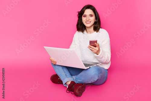 Portrait of smiling young woman wears white sweater, fashion jeans and claret shoes. Charming girl sits with crossed legs, holds laptop computer and modern smathphone isolated over pink background. photo