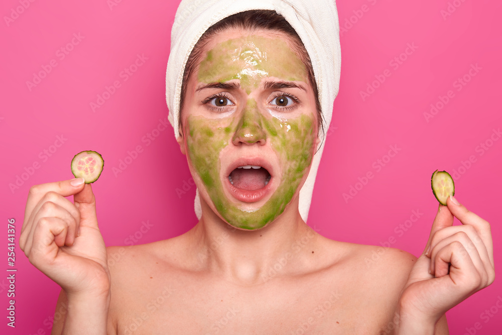Candid shot of surprised lovely young woman keeps jaw dropped, looks at herself in mirror, shocked to see green mask, applies slices of cucumber on eyes, has healthy soft skin, isolated on pink wall. Candid shot of surprised lovely young woman keeps jaw dropped, looks at herself in mirror, shocked to see green mask, applies slices of cucumber on eyes, has healthy soft skin, isolated on pink wall.