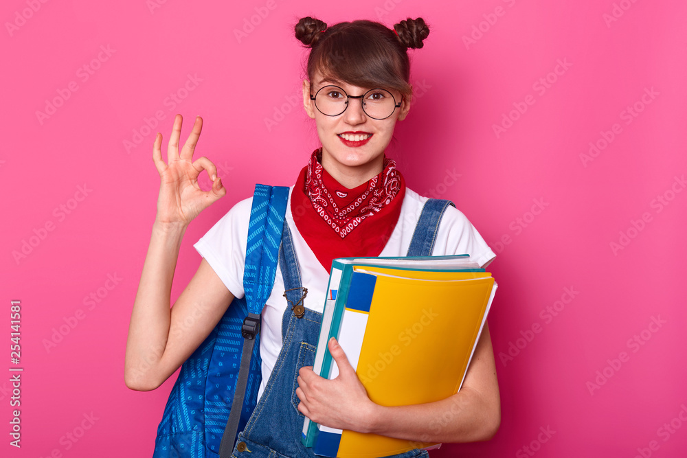 Young student showing okay sign, has good result of exam. Schoolgirl stands isolated over pink background. Happy girl studies at school. Lady holds paper folders in hands. Education concept. Young student showing okay sign, has good result of exam. Schoolgirl stands isolated over pink background. Happy girl studies at school. Lady holds paper folders in hands. Education concept.