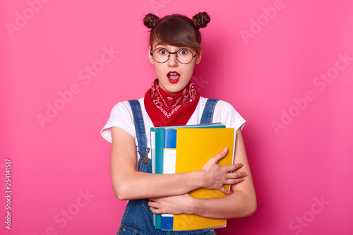 Portrait of good looking attractive girl emblaces two colurful folders for papers. Teen looks surprised, stands with opened mouth. Young lady wears white thirt, denim overalls, has funny buns on head. photo