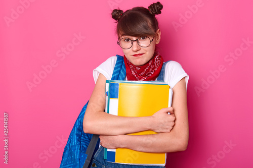 Charming student shrugs shoulders, she does not know answer friend question about new timetable. Beautiful schoolgirl holds paper folders and bag isolated over pink background. Education concept. photo