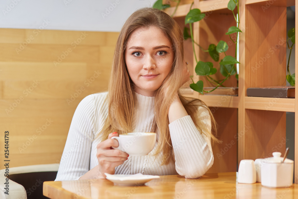 Photo of beautiful young female of European appearance, dressed in white clothes, drinks aromatic hot coffee or tea in cosy cafeteria, looks directly at camera, has good rest during weekend. Photo of beautiful young female of European appearance, dressed in white clothes, drinks aromatic hot coffee or tea in cosy cafeteria, looks directly at camera, has good rest during weekend.