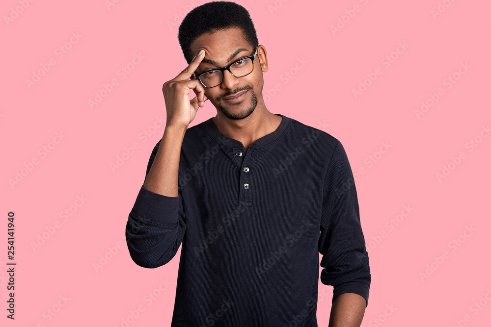 Dark skinned handsome male looks thoughtful. Man wears glasses and black sweater. Young guy with beard keeps fore finger on forehead. Model poses in photo studio isolated over rose background. Dark skinned handsome male looks thoughtful. Man wears glasses and black sweater. Young guy with beard keeps fore finger on forehead. Model poses in photo studio isolated over rose background.