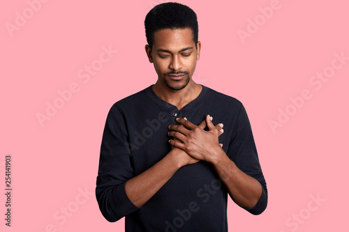 Grateful calm african man holds hands on chest, feels love, gratitude, honesty. Dark skinned guy thanks isolated on pink studio background. Female with closed eyes dresses black casual shirt. photo