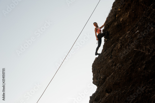 Sporty girl equipped with a rope climbing on the sloping rock and looking down