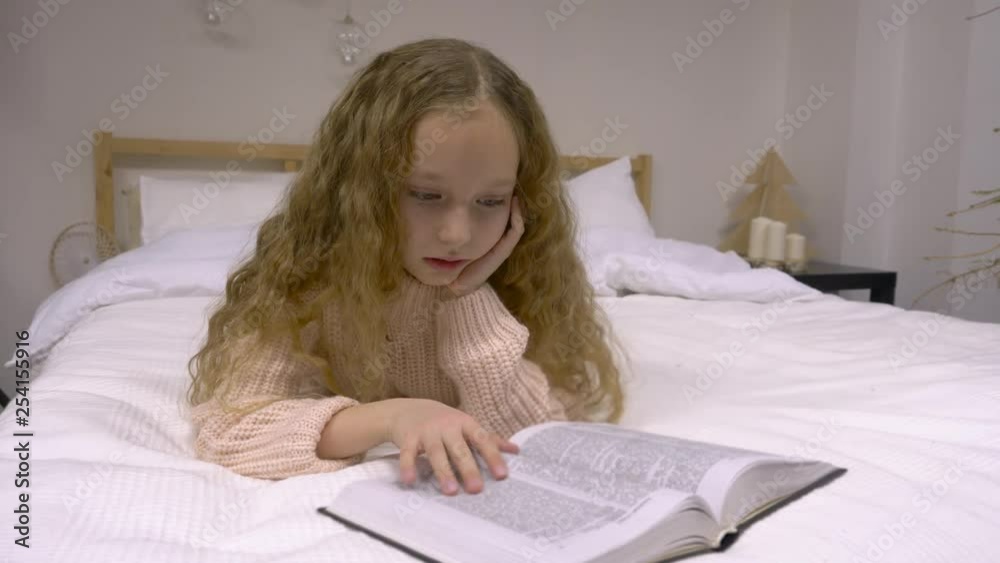 Little girl reading a book. Cute lonely little girl, teenager, reading a book while lying on the bed. Caucasian schoolgirl attentive reading an interesting book. Close up view Little girl reading a book. Cute lonely little girl, teenager, reading a book while lying on the bed. Caucasian schoolgirl attentive reading an interesting book. Close up view