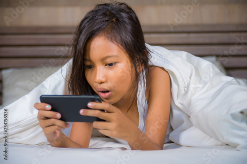 happy and beautiful 7 years old child having fun playing internet game with mobile phone lying on bed cheerful and excited in kid and domestic technology concept