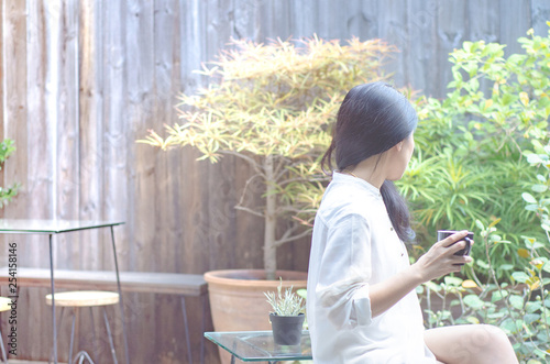 Asian women wear white shirts. She is drinking coffee in the morning garden.Black coffee mug in hand.In the garden there is a girl sitting.Do not focus on objects..
