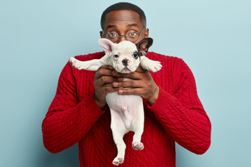 Indoor shot of dark skinned guy carries small funny puppy in front, covers face with dog, presents pet to girlfriend, likes animals, defends its rights, wears red jumper, stand over blue background
