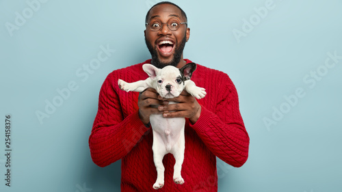 Happy man holds little puppy, rejoices it put on weight after giving dog food, satisfied after grooming procedure in salon, sleeps after bathing, hygienic care, isolated over blue background