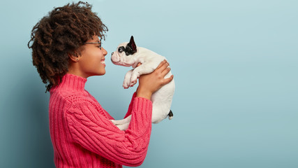 Photo of cheerful loving female stands sideways, holds little white puppy near face, teases and wants to kiss, has spare time, demonstrates good care, isolated over blue wall with copy space