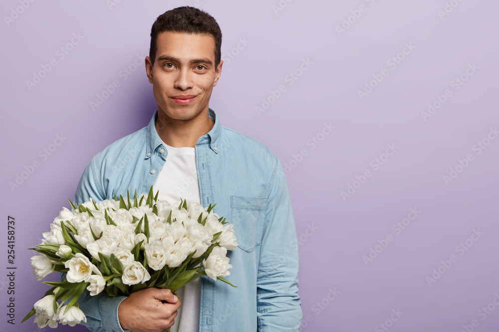 Handsome boy with bouquet of white flowers waits for girlfriend, going to make proposal and confess in love, has appealing look and gentle smile, isolated over purple studio wall, free space Handsome boy with bouquet of white flowers waits for girlfriend, going to make proposal and confess in love, has appealing look and gentle smile, isolated over purple studio wall, free space