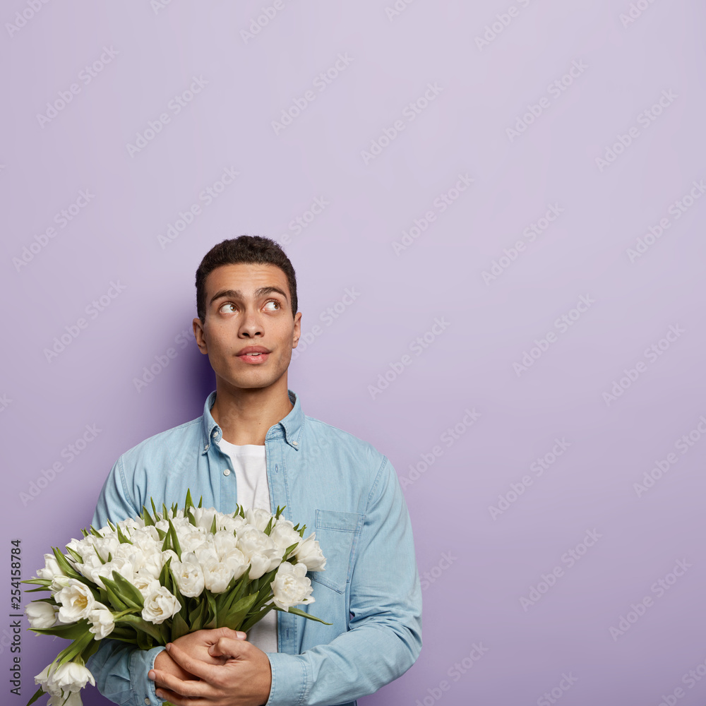 Vertical shot of thoughtful guy keeps gaze upwards, thinks about which words to say to girlfriend, holds white tulips, waits for wife returning from home, stands against purple wall with free space Vertical shot of thoughtful guy keeps gaze upwards, thinks about which words to say to girlfriend, holds white tulips, waits for wife returning from home, stands against purple wall with free space