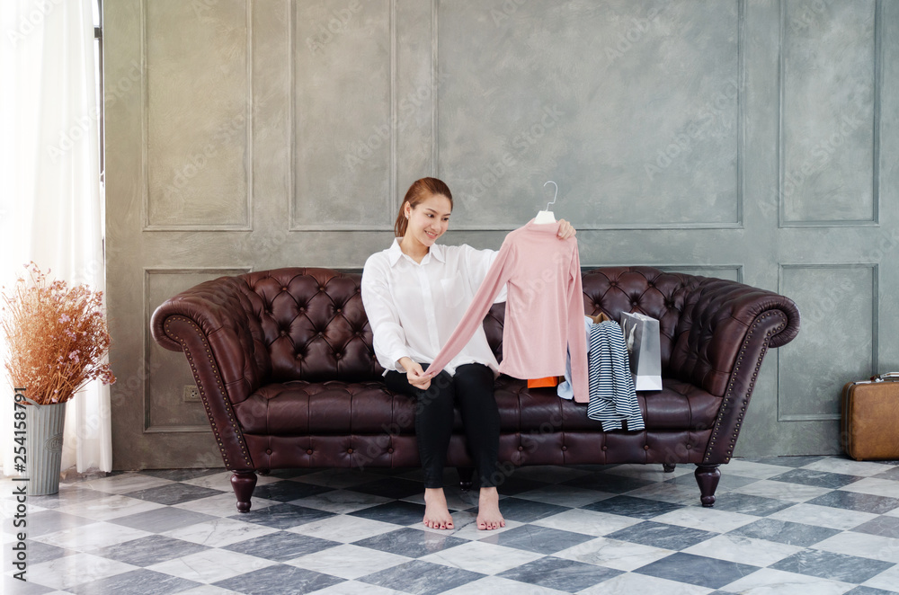 Obraz premium Beautiful Asian woman sitting on the couch.The pink shirt is in his hand.Many shirts And several paper bags on the brown sofa in the room.Woman smiling.Do not focus on the object....