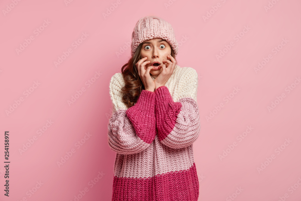 Nervous frightened woman keeps hands on face, wears hat and winter oversized jumper, worries as notices thief, expresses great disbelief, isolated over rosy background. Facial expressions concept Nervous frightened woman keeps hands on face, wears hat and winter oversized jumper, worries as notices thief, expresses great disbelief, isolated over rosy background. Facial expressions concept